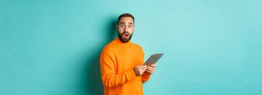 Turuncu süveter giyen, dijital tablet kullanan, açık mavi arka planda duran erkek manken görüntüsü.