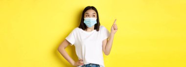 Covid, sağlık ve salgın hastalık konsepti. Tıbbi maskeli ve beyaz tişörtlü Asyalı kadın sol üst köşedeki logoyu işaret ediyor..