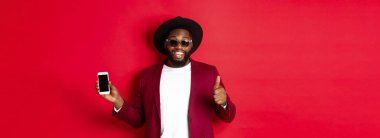 Yakışıklı ve şık siyahi adam telefon ekranı ve başparmağını kameraya gösteriyor, online mağaza uygulaması öneriyor.