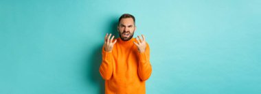 Hüsrana uğramış ve kızgın görünen öfkeli bir adamın görüntüsü, el sıkışıyor ve küçümseyerek yüzünü buruşturuyor, açık mavi arka planda sıkıntılı bir şekilde duruyor..