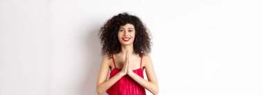 Kırmızı dudaklı çekici bir genç kadın, gece elbisesi, teşekkür ediyor ve gülümsüyor, namaste jesti ile minnettar görünüyor, beyaz arka planda duruyor..