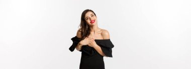 Moda ve güzellik. Siyah elbiseli çekici kadın teşekkür ediyor, gülümsüyor ve mutlu duygularla el ele tutuşuyor, beyaz arka plan..