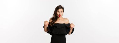 Moda ve güzellik. Siyah elbiseli zarif bir kadın, parmakları aşağıda, tanıtım yapıyor ve gülümsüyor, beyaz arka planda duruyor..