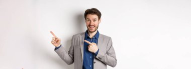 Takım elbiseli mutlu işadamı parmaklarını sağa ve gülümsüyor. Heyecanlı, özel bir anlaşma yapıyor, beyaz arka planda duruyor..