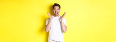 Şaşırmış ve mutlu bir adam anonslara tepki veriyor, gülümsüyor ve şaşırmış görünüyor, sarı arka planda duruyor..