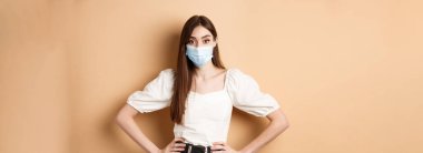 Covid-19 ve yaşam tarzı konsepti. Tıbbi maskeli kadın kameraya bakıyor, bej arka planda duruyor..