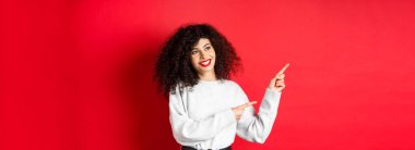 Kıvırcık saçlı ve makyajlı güzel bir Avrupalı kadın, parmaklarını sola uzatıyor ve rüya gibi bir gülümsemeyle yan yana bakıyor, terfi anlaşmasına bakıyor, kırmızı arka plan..