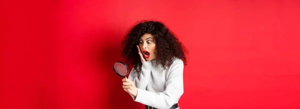 Shocked girl look through magnifying glass with dropped jaw, seeing something amazing, standing on red background.