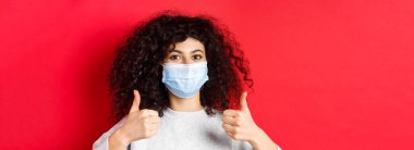 Sağlıklı insanlar ve covid-19 konsepti. Kıvırcık saçlı, salgın sırasında kendini korumak için tıbbi maske takan gülümseyen kadının yakın plan portresi..
