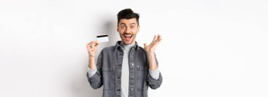 Heyecanlı adam sevinçten çığlık atıyor ve plastik kredi kartı gösteriyor, alışveriş yaparken seviniyor, beyaz arka planda duruyor..