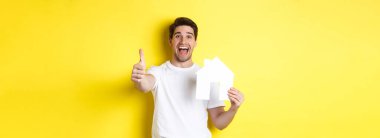 Emlak konsepti. Mutlu genç bir erkek alıcı, başparmağını gösteriyor ve kağıt ev modeli, gülümsüyor, sarı arka planda duruyor..