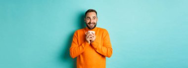 Mutlu genç adam kahve içiyor, gülümsüyor ve rüya gibi bir yüzle soluna bakıyor, turkuaz arka planda dikiliyor..