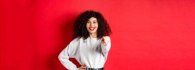 Kıvırcık saçlı, neşeli bir kadın, kamerayı işaret ediyor, seni davet ediyor, birini seçiyor, kırmızı arka planda duruyor..