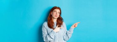 Sinirli, genç, kızıl saçlı kız gözlerini yuvarlıyor, parmaklarını sıkıcı ya da topal bir şeye doğrultuyor, mavi arka planda tedirgin bir şekilde duruyor..