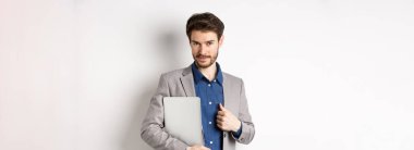 Başarılı bir iş adamı şık bir takım elbise içinde dizüstü bilgisayar taşıyor ve kameraya kendinden emin bakıyor, gülümsüyor memnun, beyaz bir altyapısı var..