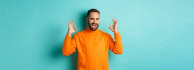 Kendine güveni tam bir adam, tamam işaretleri gösteriyor ve sana göz kırpıyor, ürün öneriyor, stüdyo arka planında turuncu süveter giyiyor..