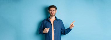 Hüsrana uğramış ve şüpheci adam somurtuyor, dudaklarını büzüyor ve parmaklarını boş yere doğrultuyor, kötü şeyden yakınıyor, mavi arka planda üzgün duruyor..