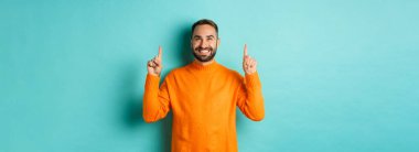 Memnun ve mutlu alıcı parmaklarını havaya kaldırarak gülümsüyor turkuaz arka planda turuncu süveter giyiyor..