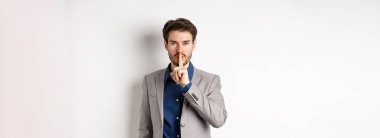 Yakışıklı maço takım elbiseli adam kameraya şşş yapıyor, parmak dudağa basıyor, sesini alçalt, sırrını söyle, beyaz arka plana karşı duruyor..