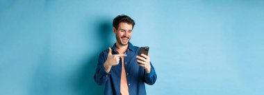 Mobil ekranı okuyan, telefonu işaret eden, online tanıtım ya da akıllı telefon uygulamasını kontrol eden, mavi arka planda duran yakışıklı bir adam resmi.