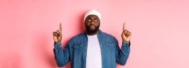 Endişeli ve tereddütlü siyah adam, bereli parmaklarını havaya kaldırarak, şüpheli yüzüyle, pembe arka planda dikiliyor..