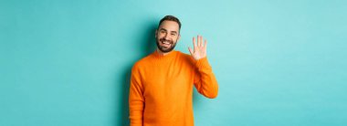 Dost canlısı genç adamın merhaba derken fotoğrafı, gülümseyen ve feragat eden el, seni selamlayan, açık mavi arka planda turuncu süveter giymiş..