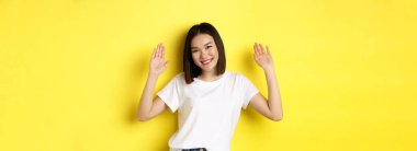 Dost canlısı genç Asyalı kadın merhaba diyor, boş ellerini kaldırıyor ve gülümsüyor, seni selamlıyor, sarı arka planda duruyor..