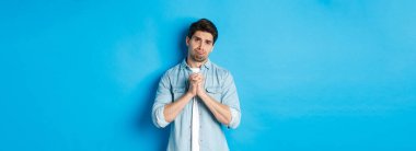 Yardım için yalvaran, somurtan ve üzgün görünen genç bir adam bir şey istiyor, mavi arka plana karşı duruyor..