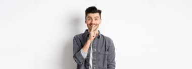 Mutlu adam kameraya şşşş yapıyor, parmakları dudaklarında gülümsüyor, sessiz kalmak istiyor, büyük bir sır veriyor ya da sürpriz yapıyor, beyaz arka planda duruyor..