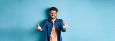 Angry caucasian man say fuck you and showing middle fingers, make rude gesture with pissed-off face, standing annoyed on blue background.