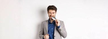 Başarılı takım elbiseli genç işadamı kredi kartını ısırıyor ve beyaz arka planda arsız görünüyor..