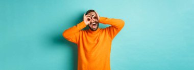 Komik ve mutlu bir adamın el ele tutuşup sahte gözlüklerden bakıp güldüğü turkuaz arka planda turuncu süveter giydiği görüntüsü..