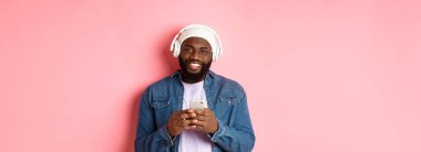 Yakışıklı siyahi adamın kulaklıklı, müzik dinleyen ve cep telefonu kullanan, kameraya gülümseyen, pembe arka planlı bir resmi..