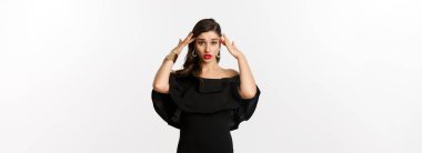 Moda ve güzellik. Siyah elbiseli, kızgın ve yorgun bir kadın. Başa dokunuyor ve yuvarlanıyor. Beyaz arka planda sıkıntılı duruyor..