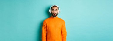 Kulaklıkla müzik dinleyen, sesten etkilenmiş kameraya bakan, turkuaz arka planda duran yetişkin bir adam..