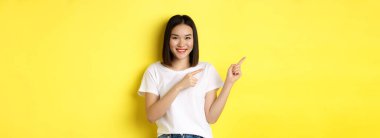 Güzellik ve moda konsepti. Beyaz tişörtlü güzel Asyalı kadın, sarı arka planda durup, parmaklarını sola gösteriyor..