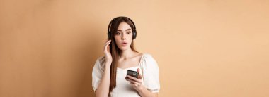 Harika melodiler. Heyecanlı genç bir kız yeni şarkıya hayran, kulaklıkla müzik dinliyor ve etkilenmiş görünüyor, elinde cep telefonu, bej arka plan.