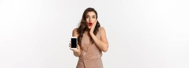 İnternetten alışveriş. Lüks elbiseli zarif bir kadın akıllı telefon ekranı gösteriyor, kameraya hayran bakıyor, beyaz arka plan.