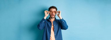 Yakışıklı beyaz bir adamın gözlük takıp kameraya baktığı, gözlükçüde yeni gözlük denediği, mavi arka plan..