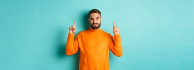 Şüpheci ve gönülsüz bir adam hoşnutsuz görünüyor, parmakla işaret ediyor, kötü bir teklif, turkuaz arka planda duruyor..