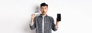 Plastik kredi kartı ve cep telefonu ekranıyla beyaz arka planda duran heyecanlı yakışıklı adam..