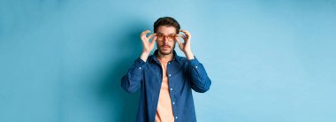 Yakışıklı genç adam mağazada yeni gözlük deniyor, kameraya bakıyor, mavi arka planda duruyor..