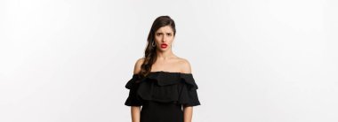 Moda ve güzellik. Siyah elbiseli, hayal kırıklığına uğramış ve üzgün bir kadın kameraya hoşnutsuzca bakıyor, kıskanç bakışlarla şikayet ediyor, beyaz arka planda duruyor..