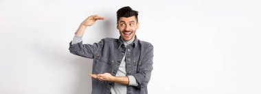 Heyecanlı adam logoya bakıyor ve büyük bir şey gösteriyor, büyük bir nesne şekillendiriyor ve şaşkın bir şekilde gülümsüyor, beyaz arka planda kot ceketle duruyor..