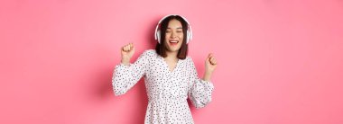 Mutlu genç Asyalı kadın dans ediyor ve eğleniyor, kulaklıkla müzik dinliyor, pembe arka planda duruyor. Boşluğu kopyala