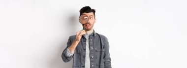 Komik genç adam gözlerini kısıyor, büyüteçle yüzlerini gösteriyor, beyaz arka planda duruyor..