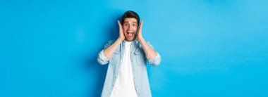 Korkmuş bir adam çığlık atıyor ve bir şeye bakıyor, mavi arka planda duruyor..