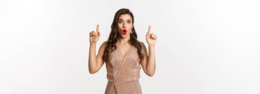 Yeni yıl, Noel ve kutlama konsepti. Şaşırmış ve heycanlı bir kadın, parti için kıyafet giyiyor, parmakla işaret ediyor, tanıtım teklifini gösteriyor, beyaz arka plan.