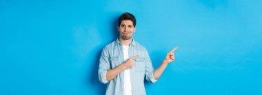 Şüpheci yetişkin adam parmaklarını sol tarafı işaret ediyor, şüpheli ve kararsız görünüyor, mavi arka planda duruyor..