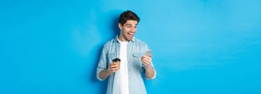 Akıllı telefona şaşırmış bakan, kahve içen, mavi arka planda duran adam şaşırmış..